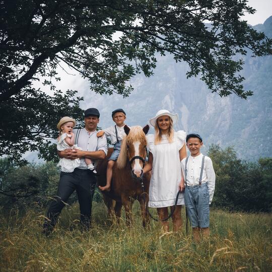 En familie på fem kledd i historiske klær. De står i gresset ved et tre sammen med en hest.