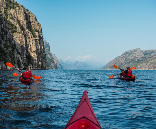 3 red kayaks in the blue fjord paddling with mountains surrounding them. Blue skies.
