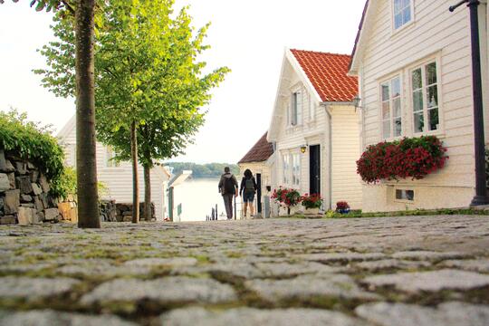 Bilde av hvite trehus i gamle Stavanger med to mennesker som spaserer på brosteinen i bakgrunnen av bildet.