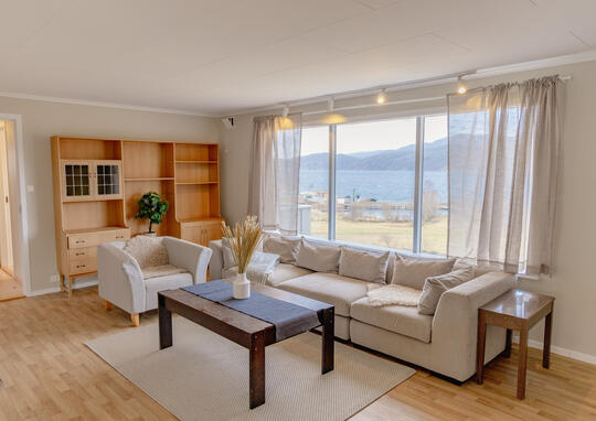 A living room in a hoilday home. Decorated in cream and white in a nordic style. Views of the fjord out the window. The photo shows a sofa, a chair, a coffee table and a book case. Holiday house near Lauvvik