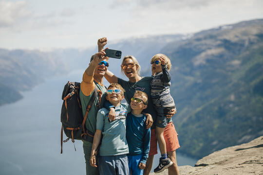 Familie på fem tar selfie på toppen av et fjell med fjord og fjell som bakgrunn