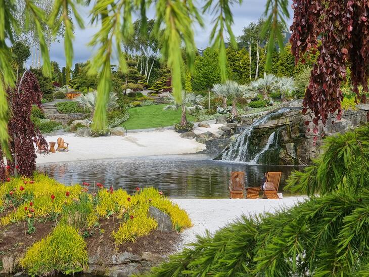 En tropisk hage med fargerike planter, et lite vann med foss omkranset av hvit sand og lounge stoler, på Flor & Fjære