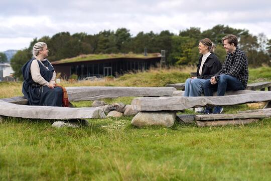 Three people sitting on log benches outside the iron age farm. The lady of the house talks to her guest.