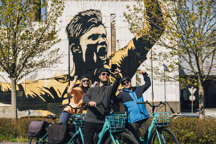 Three people on a bike take a selfie in front of a mural