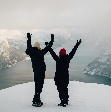To personer med hendene i været på en fjelltopp med utsikt til fjord i snø